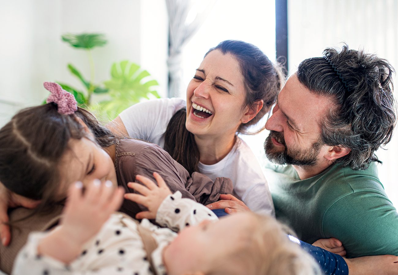 A happy family of four, including two adults and two young children, laughing and playing together indoors, creating a joyful and loving atmosphere.