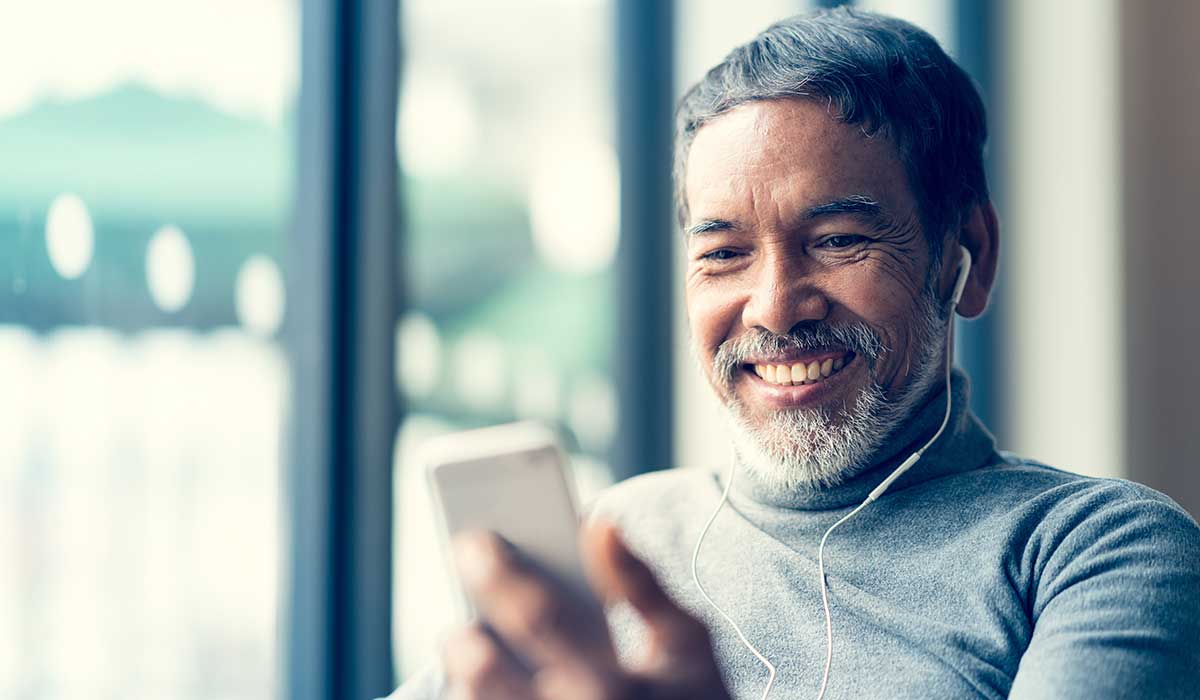 Smiling middle-aged man with gray hair and beard, wearing a turtleneck and earphones, looking at his smartphone while sitting by a window.