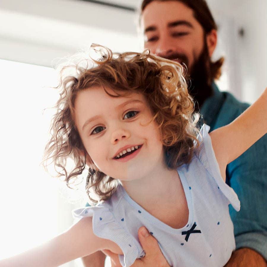 A smiling young child with curly hair is held up playfully by an adult with a beard in a bright, sunlit room. The child has arms outstretched and looks happy.